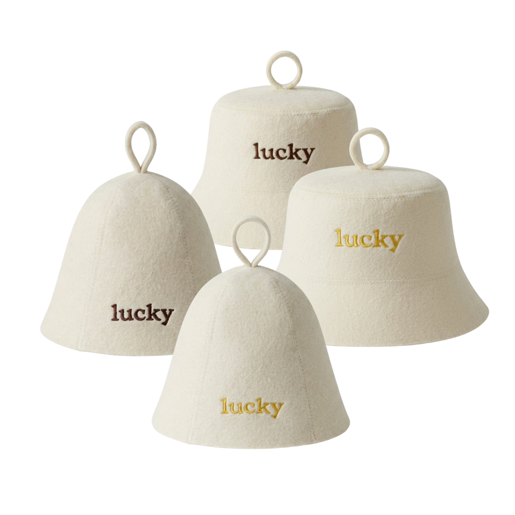 Four cream-colored felt bell-shaped sauna hats with hanging loops, each embroidered with the word 'lucky' in different font colors: two in dark brown, two in gold.